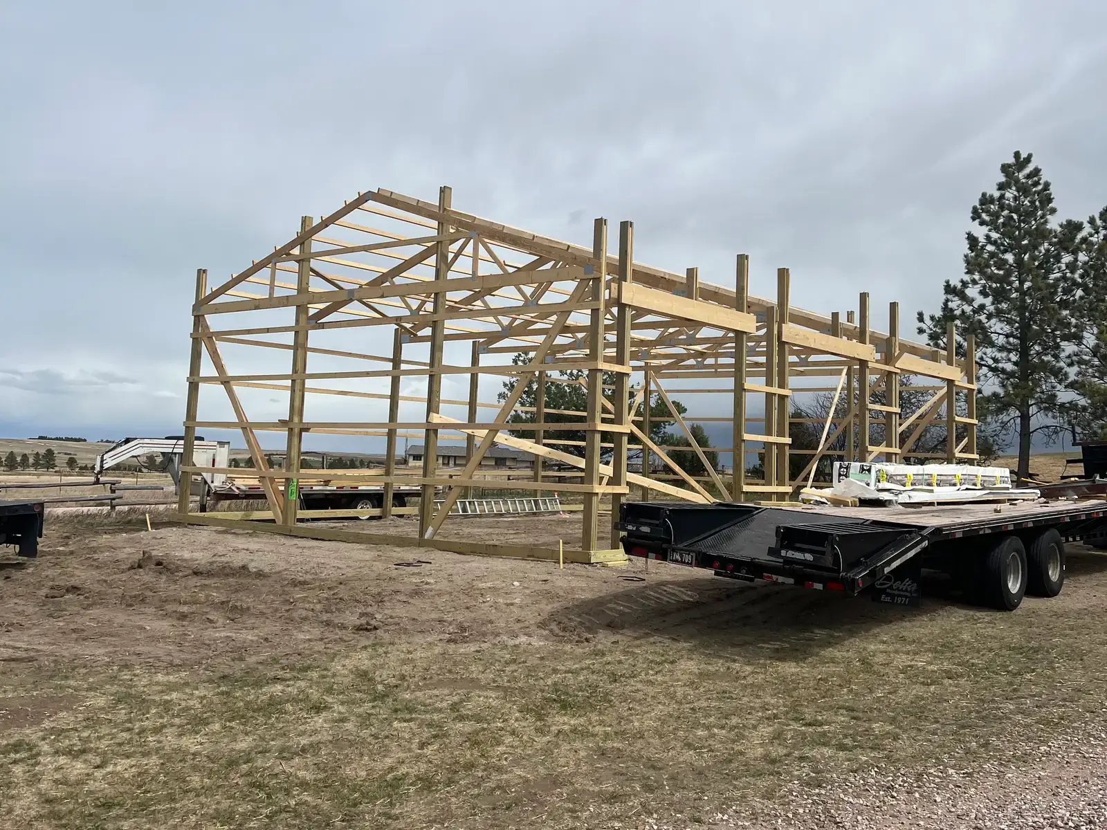 Open post-frame garage structure with roof trusses, wall framing and a material trailer alongside.